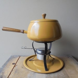 May include: Mustard yellow fondue pot with a wooden handle and lid. The pot sits on a black metal stand with a burner and a matching yellow base. A vintage kitchenware item.