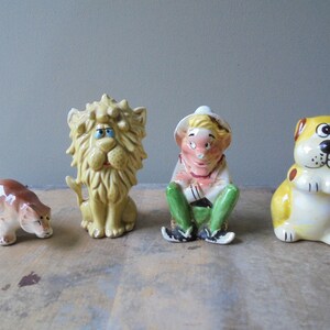 May include: Four ceramic figurines: a brown bear, a yellow lion, a seated figure in a hat, and a yellow dog. The lion has a yellow mane and blue eyes. The seated figure wears a hat, pink shirt, and green pants. The dog has brown spots.