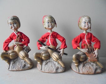 May include: Three vintage figurines of elves wearing red and gold clothing. Each elf is playing a different musical instrument: a flute, a harp, and a tambourine.
