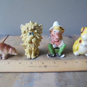 May include: Four vintage ceramic figurines: a brown bear, a yellow lion, a seated figure in a hat, and a yellow dog with brown spots. The figurines are arranged on a wooden surface with a ruler.