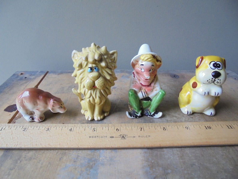 May include: Four vintage ceramic figurines: a brown bear, a yellow lion, a seated figure in a hat, and a yellow dog with brown spots. The figurines are arranged on a wooden surface with a ruler.