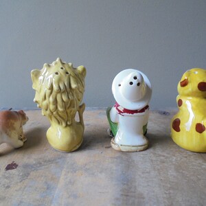 May include: A collection of four vintage ceramic salt and pepper shakers. The set includes a bear, a lion, a cowboy, and a giraffe. The shakers are painted in various colors, including yellow, brown, and white. The lion and cowboy shakers have holes for dispensing.