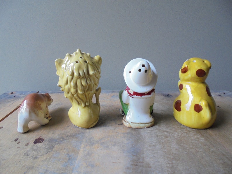 May include: A collection of four vintage ceramic salt and pepper shakers. The set includes a bear, a lion, a cowboy, and a giraffe. The shakers are painted in various colors, including yellow, brown, and white. The lion and cowboy shakers have holes for dispensing.