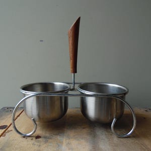 May include: A vintage stainless steel condiment holder with two bowls and a wooden handle. The holder has a curved metal frame and is designed to hold salt, pepper, or other spices. The wooden handle is a dark brown color.