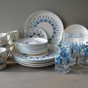 May include: A collection of white ceramic dinnerware and clear glass drinkware with blue and green floral designs. The set includes plates, bowls, cups, and glasses, all with a matching aesthetic. The plates and bowls have a blue trim.