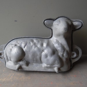 May include: A vintage, silver-colored lamb-shaped cake mold. The mold is made of metal and features a detailed design of a lamb with its head turned slightly. It has handles on the sides and small feet. The lamb mold is likely used for baking Easter cakes.