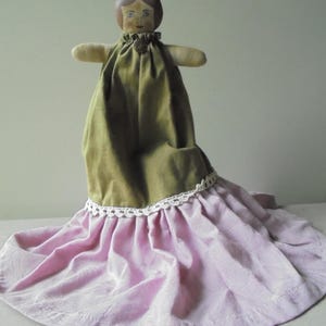 K&ouml;nnte beinhalten: Eine handgemachte Stoffpuppe mit bemalten Gesichtsz&uuml;gen, die ein gr&uuml;nes Kleid und einen rosa Rock mit wei&szlig;er Spitzenborte tr&auml;gt.