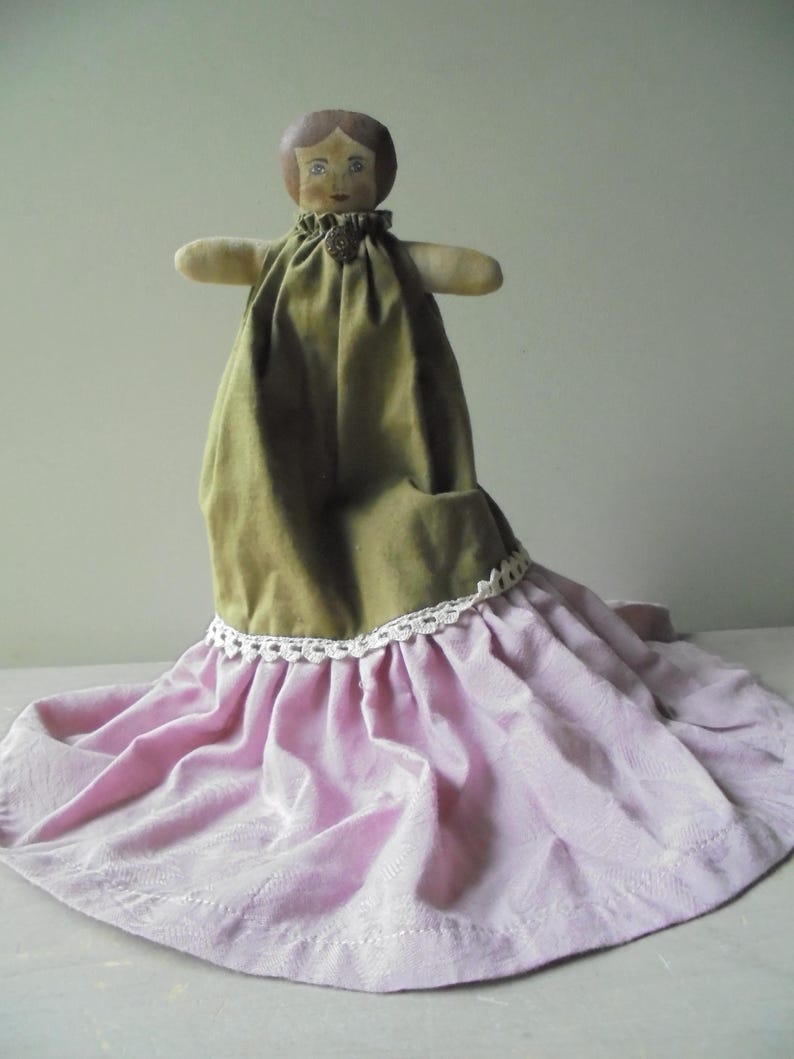 K&ouml;nnte beinhalten: Eine handgemachte Stoffpuppe mit bemalten Gesichtsz&uuml;gen, die ein gr&uuml;nes Kleid und einen rosa Rock mit wei&szlig;er Spitzenborte tr&auml;gt.