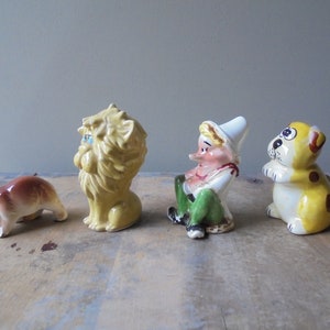 May include: A collection of four ceramic figurines. A brown bear, a yellow lion with blue eyes, a seated elf with a white hat and green pants, and a yellow dog with brown spots. The figurines are arranged on a wooden surface.