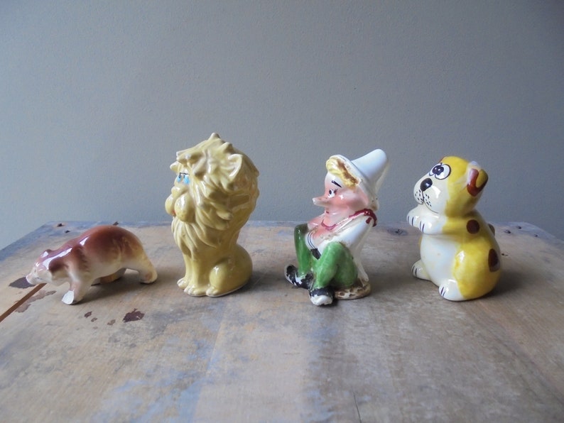 May include: A collection of four ceramic figurines. A brown bear, a yellow lion with blue eyes, a seated elf with a white hat and green pants, and a yellow dog with brown spots. The figurines are arranged on a wooden surface.