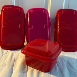 May include: A set of four plastic food storage containers with lids. Three containers are rectangular with red and magenta lids positioned vertically, while one square red container with a matching lid sits in the foreground.