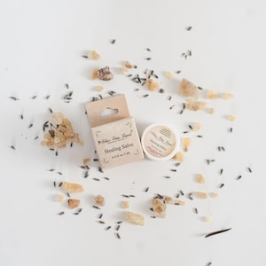May include: A small white jar with a brown label that reads "Relax, Renew, Repeat" and "Healing Salve" with a measurement of 0.25 fl oz (7 ml). The jar is surrounded by small pieces of dried lavender and frankincense.