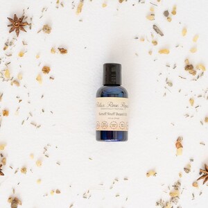 May include: A blue bottle of beard oil with a white label that reads "Relax Rinse Repeat" and "Scruff Stuff Beard Oil" with a list of ingredients. The bottle is surrounded by dried herbs and spices on a white background.