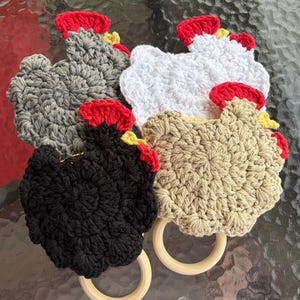 May include: Four crochet chicken-shaped ornaments, each featuring a bright red comb and a small yellow beak. The chickens are crafted in gray, white, black, and beige yarn. Two of the figures are attached to smooth, round, light-colored wooden rings.