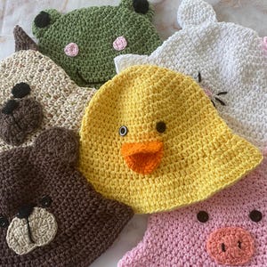 May include: A collection of handmade crochet hats in various animal designs. The hats include a yellow duck, a green frog, a brown bear, a white bunny, a tan bear, and a pink pig. Each hat features detailed facial features.