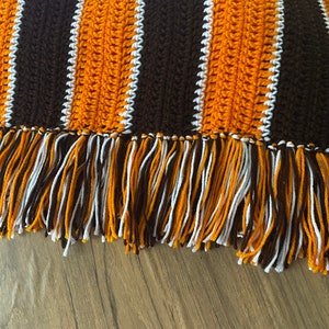 May include: A crocheted throw blanket with brown, orange, and white stripes and a fringe border.