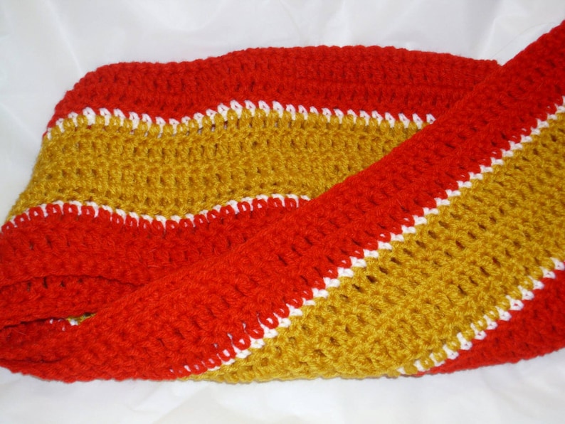 May include: A crocheted scarf with red, white, and gold stripes. The scarf is made with a simple stitch and has a soft, textured feel.