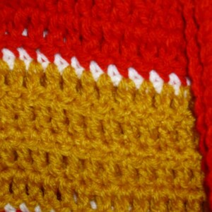 May include: Close-up of a crocheted fabric with a red, white, and gold pattern. The fabric is made of yarn and has a textured surface.