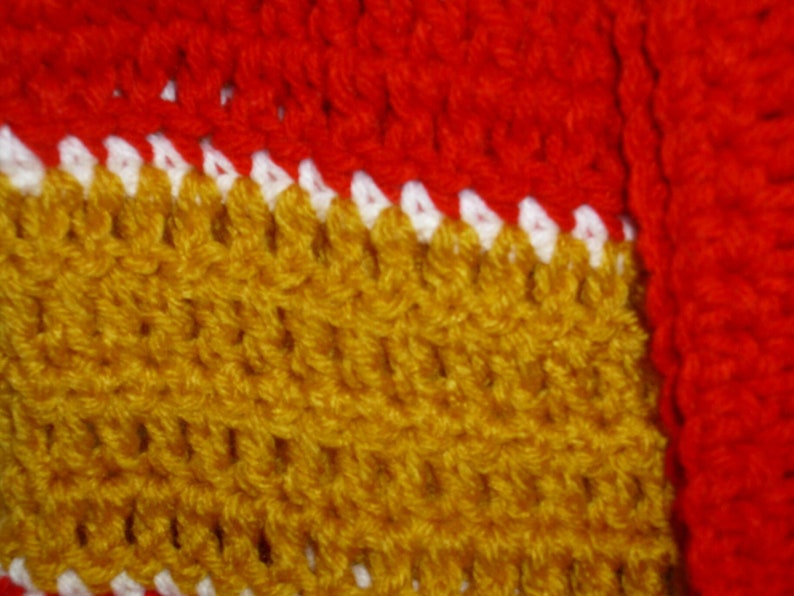 May include: Close-up of a crocheted fabric with a red, white, and gold pattern. The fabric is made of yarn and has a textured surface.