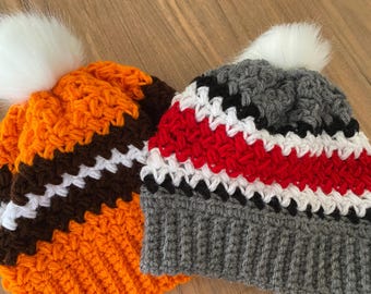 May include: Two handmade crochet beanies with white faux fur pom-poms. One beanie is orange, brown, and white. The other is gray, red, black, and white. The beanies are made with a textured stitch.