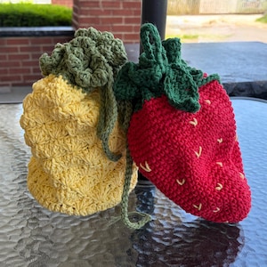 May include: Two crocheted produce bags: a yellow pineapple and a red strawberry. The pineapple bag has a green leafy top and a drawstring closure. The strawberry bag features a green leafy top and small yellow seed details.