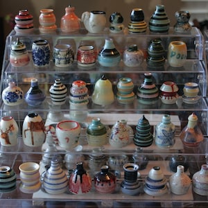 May include: A tiered display shelf filled with a collection of small, colorful ceramic vases. The vases are decorated with various patterns, including stripes, dots, and floral designs. Some of the vases have a glossy finish, while others have a matte finish.