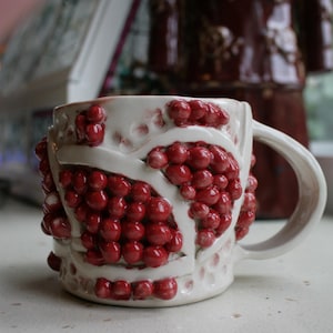 May include: A white ceramic mug featuring a textured surface with raised, glossy red berry-like clusters separated by smooth white bands. The mug has a curved handle and a dimpled, cratered texture on the base and upper rim areas.