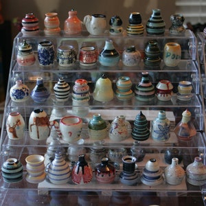 May include: A tiered display shelf filled with a variety of small, colorful ceramic vases. The vases are decorated with different patterns, including stripes, dots, and floral designs. Some of the vases have a glossy finish, while others have a matte finish.