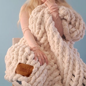 May include: A woman is sitting on a chair wrapped in a large, cream-colored chunky knit blanket. The blanket has a brown leather tag with the word "Dominick" on it.