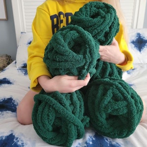 May include: A person holding four large, green, fluffy yarn balls. The yarn is soft and textured, and the balls are arranged in a pile.