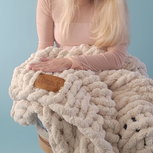 Pu&ograve; includere: Una donna &egrave; seduta su uno sgabello di legno con una grande coperta di maglia grossa di colore crema. La coperta ha un'etichetta in pelle marrone con il nome "Dominick" su di essa.
