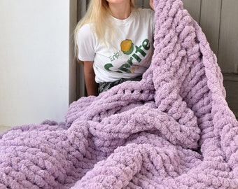 Könnte beinhalten: Eine Frau sitzt auf dem Boden mit einer großen, grob gestrickten Decke in helllila. Die Decke ist aus dickem Garn gefertigt und hat ein strukturiertes, Zopfmuster. Die Frau trägt ein weißes T-Shirt mit dem Aufdruck "Sprite".