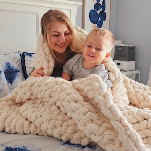 Pu&ograve; includere: Una donna e un bambino piccolo sono sdraiati sotto una grande coperta di maglia bianca e spessa su un letto con un copriletto a motivi blu e bianchi.