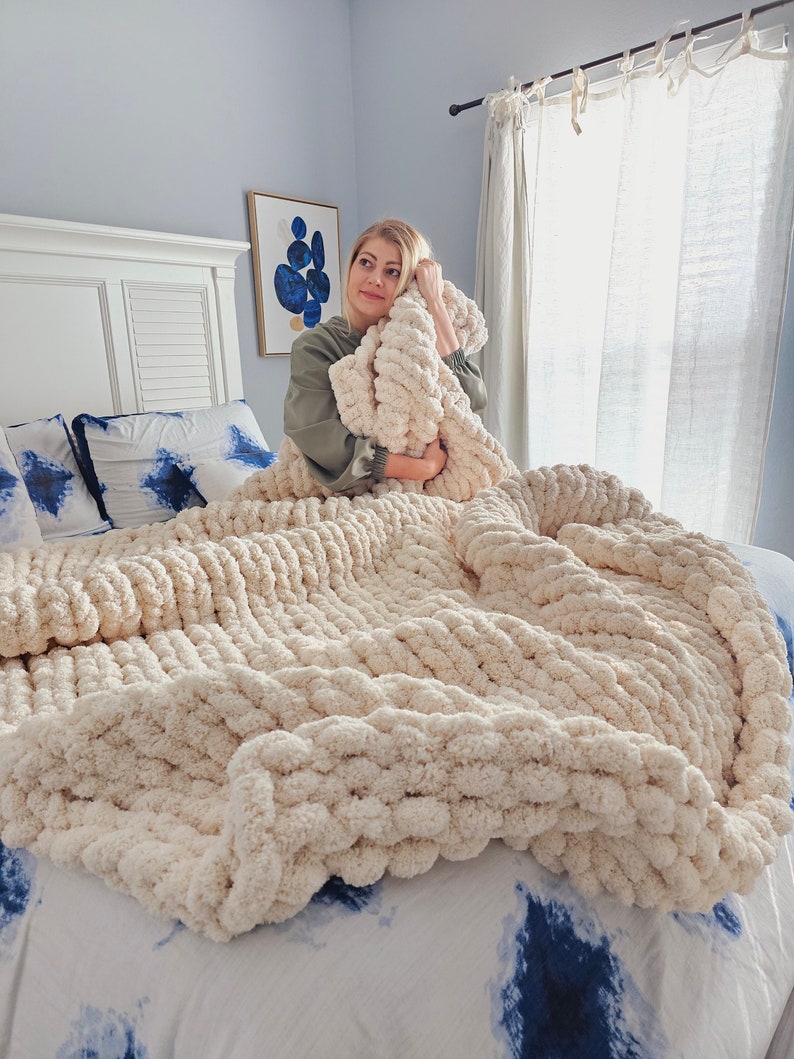 Pu&ograve; includere: Una donna &egrave; seduta su un letto con una coperta di maglia bianca spessa. La coperta &egrave; realizzata con filato spesso e ha una consistenza morbida e soffice. Il letto ha una testiera bianca e un copripiumino con motivo blu e bianco.