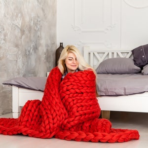 May include: A woman sits on the floor in front of a bed, wrapped in a large, chunky knit red blanket. The blanket is made of thick yarn and has a loose, open weave.