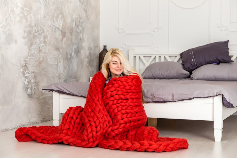 May include: A woman sits on the floor in front of a bed, wrapped in a large, chunky knit red blanket. The blanket is made of thick yarn and has a loose, open weave.