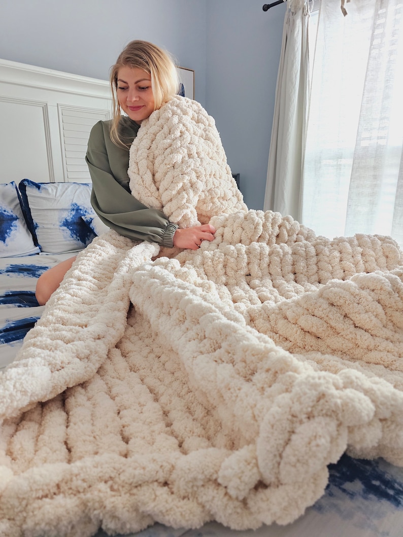 Pu&ograve; includere: Una coperta di maglia grossa color crema con un motivo a treccia. La coperta &egrave; drappeggiata su un letto con biancheria da letto a motivi blu e bianchi.