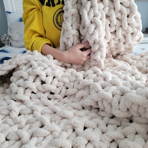 May include: A large, cream-colored chunky knit blanket with a soft, fluffy texture. The blanket is made with thick yarn and has a loose, open weave.