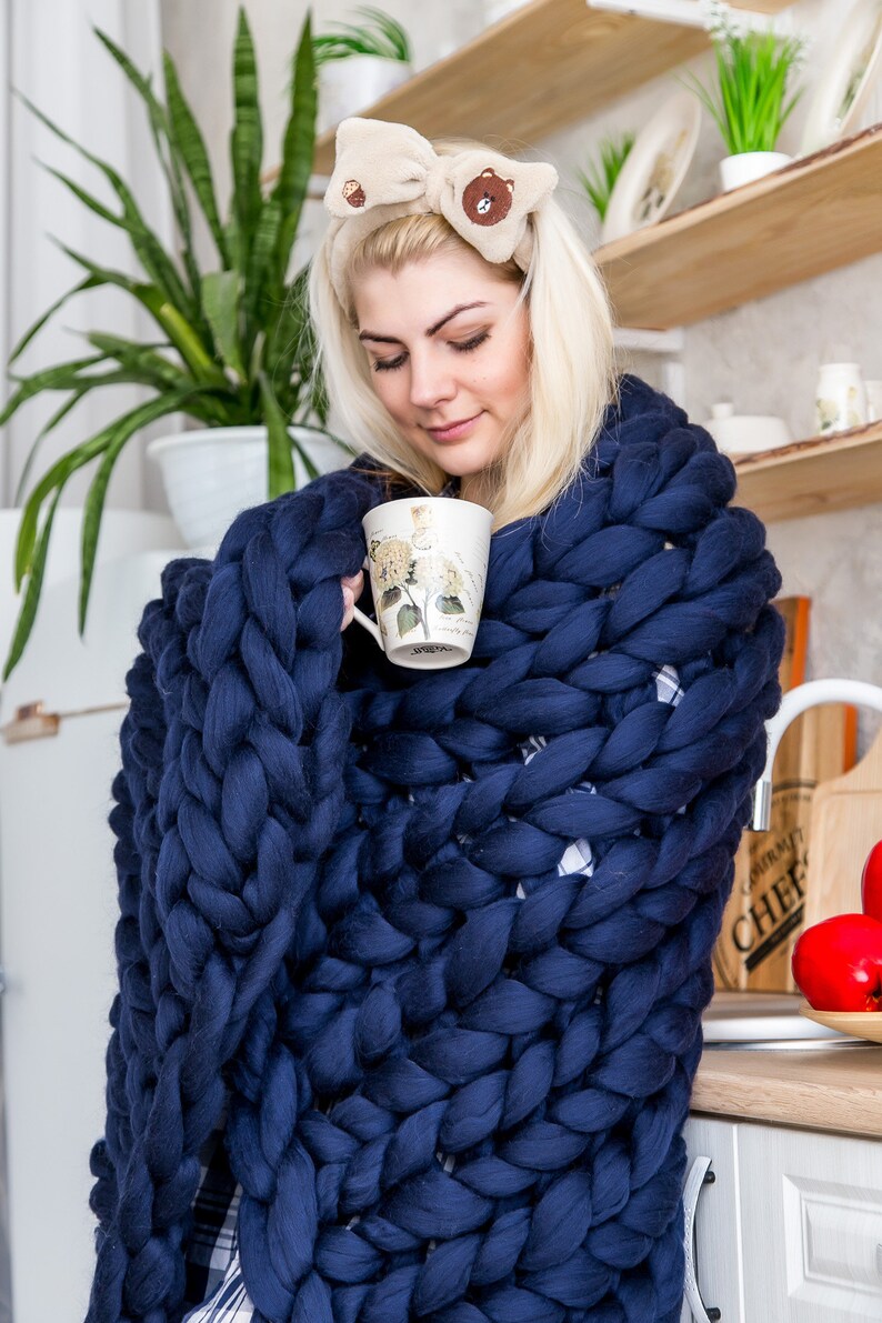 May include: A person wrapped in a navy blue chunky knit blanket, holding a white mug with floral designs. The blanket is made of thick, soft yarn and has a cozy, comforting look.