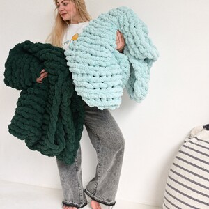 Pu&ograve; includere: Una donna tiene in mano due coperte di maglia spessa, una in turchese e una in verde scuro. Le coperte sono fatte con un filo spesso e hanno una consistenza morbida e soffice.