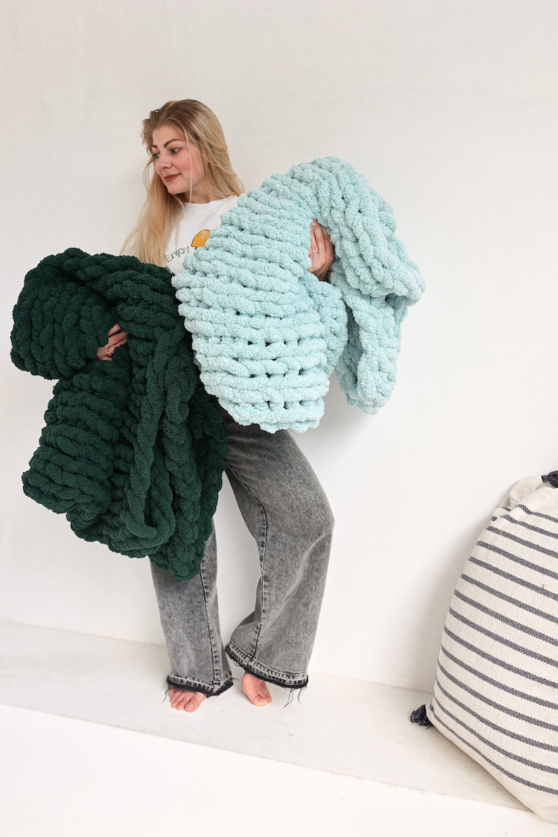 Pu&ograve; includere: Una donna tiene in mano due coperte di maglia spessa, una in turchese e una in verde scuro. Le coperte sono fatte con un filo spesso e hanno una consistenza morbida e soffice.