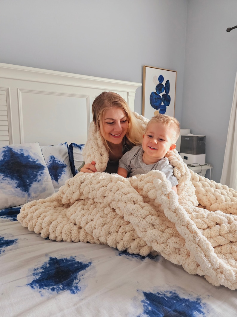 Pu&ograve; includere: Una donna e un bambino piccolo sono sdraiati sotto una grande coperta di maglia bianca e spessa su un letto con un copriletto a motivi blu e bianchi.