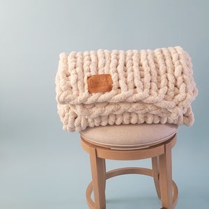 Pu&ograve; includere: Una coperta di maglia grossa di colore beige chiaro con un'etichetta in pelle marrone che dice "Dominick" &egrave; piegata e posizionata su uno sgabello di legno con una seduta in tessuto beige chiaro.