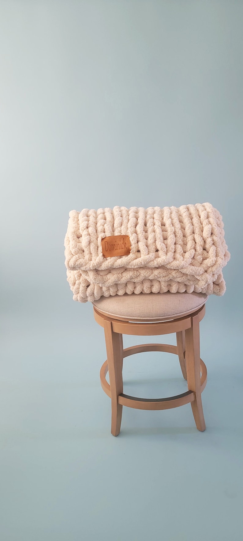 Pu&ograve; includere: Una coperta di maglia grossa di colore beige chiaro con un'etichetta in pelle marrone che dice "Dominick" &egrave; piegata e posizionata su uno sgabello di legno con una seduta in tessuto beige chiaro.