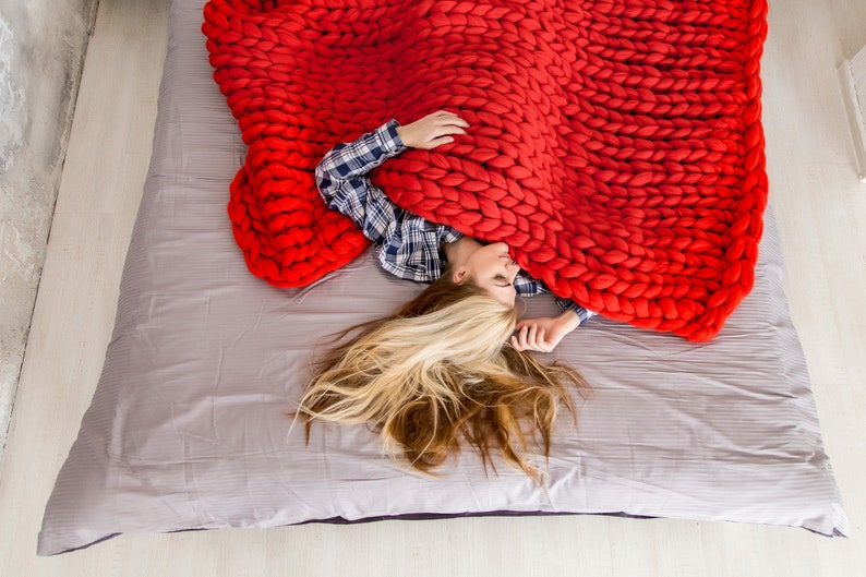 May include: A person is lying in bed under a large, chunky knit red blanket. The blanket is made of thick yarn and has a soft, cozy texture.