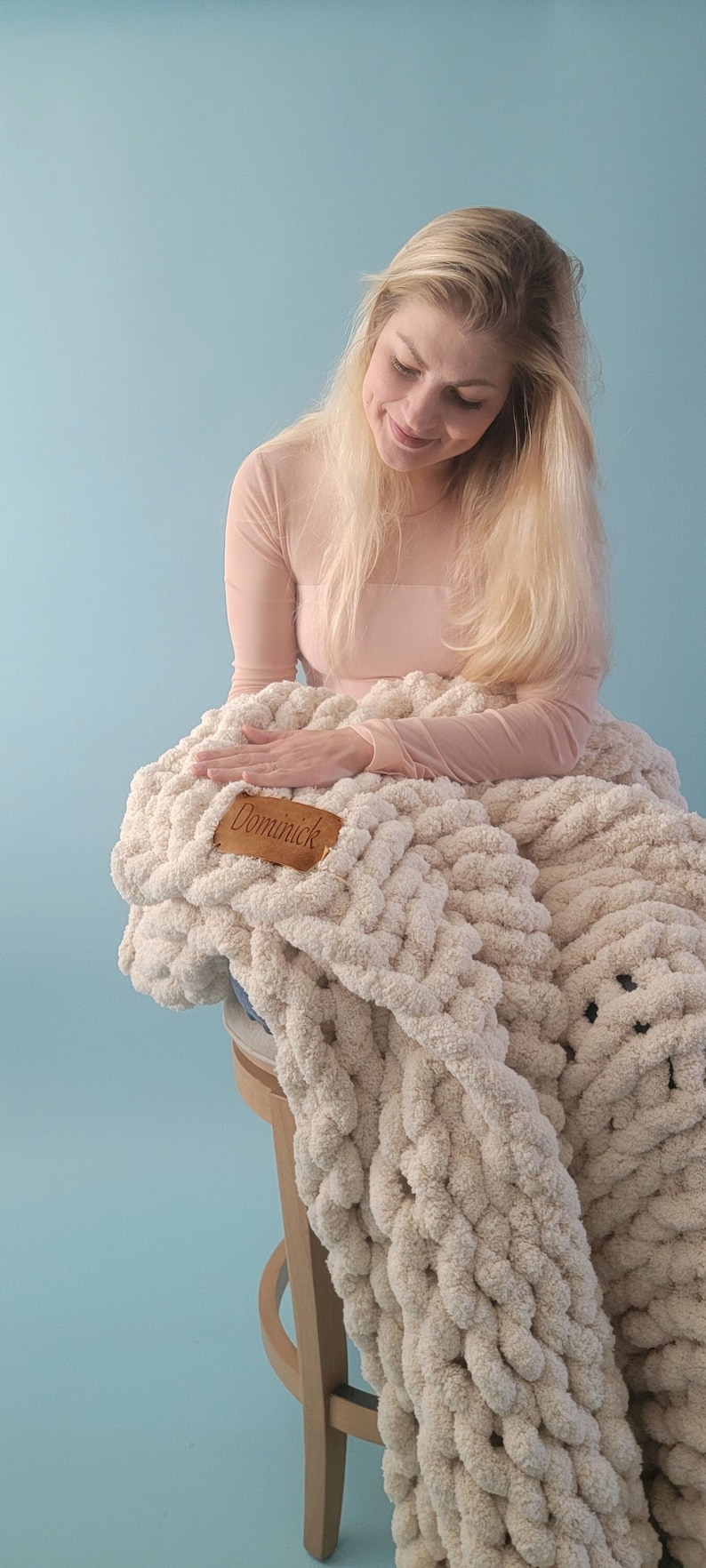 Pu&ograve; includere: Una donna &egrave; seduta su uno sgabello di legno con una grande coperta di maglia grossa di colore crema. La coperta ha un'etichetta in pelle marrone con il nome "Dominick" su di essa.