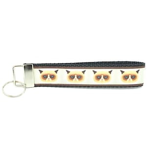 May include: A black and brown key chain with a white background featuring a repeating pattern of a grumpy cat face.