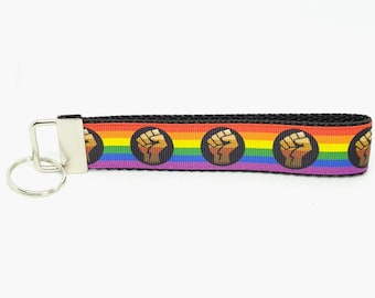 May include: A rainbow-striped keychain wristlet with a black border and a silver-colored metal clasp. The wristlet features a repeating pattern of a raised fist inside a black circle.