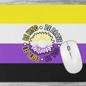May include: A mousepad featuring the non-binary pride flag with a yellow, white, purple, and black color scheme. The flag has a sunflower in the center with the text "Be Kind, Be Brave, Be True, Be You" around it.