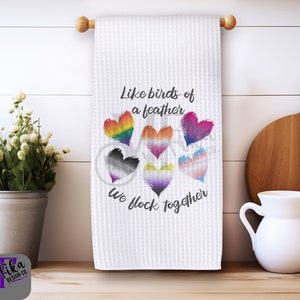 May include: White kitchen towel with a rainbow, black and white, and yellow and purple heart design. The text reads "Like birds of a feather we flock together".